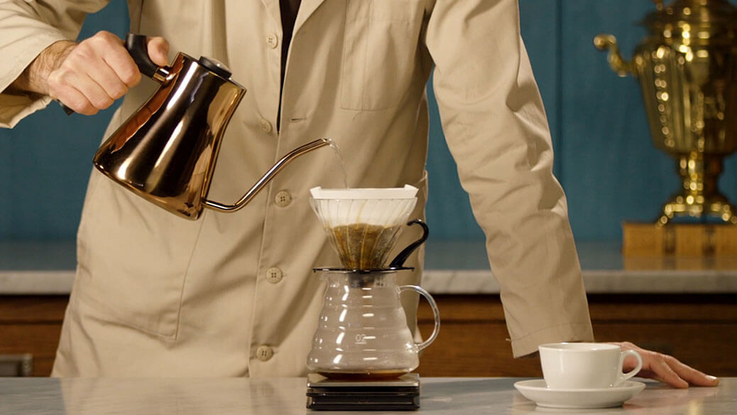 Person pouring coffee from a kettle into a pour-over coffee maker with a cup and saucer on a table.